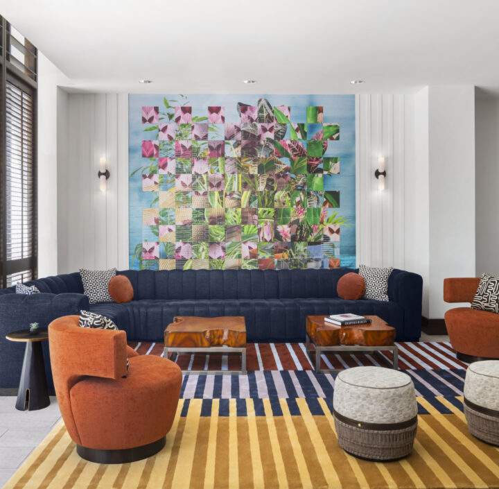 Modern hotel lobby with a blue sofa, orange chairs, striped rug, round ottomans, geometric wall art, and large window with blinds.