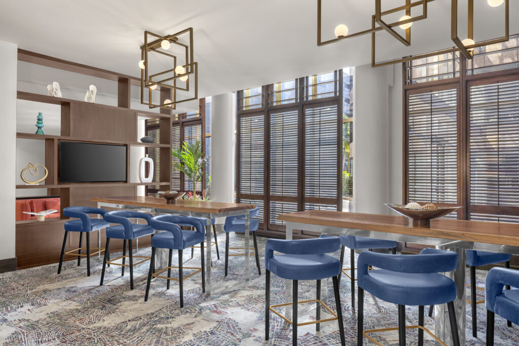 Modern lounge with two wooden tables, blue velvet chairs, large windows with blinds, abstract rug, and geometric pendant lights.