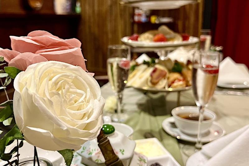 Close-up of white and pink roses on a table set for afternoon tea with champagne, tea, and a tiered tray of sandwiches and desserts in the background.