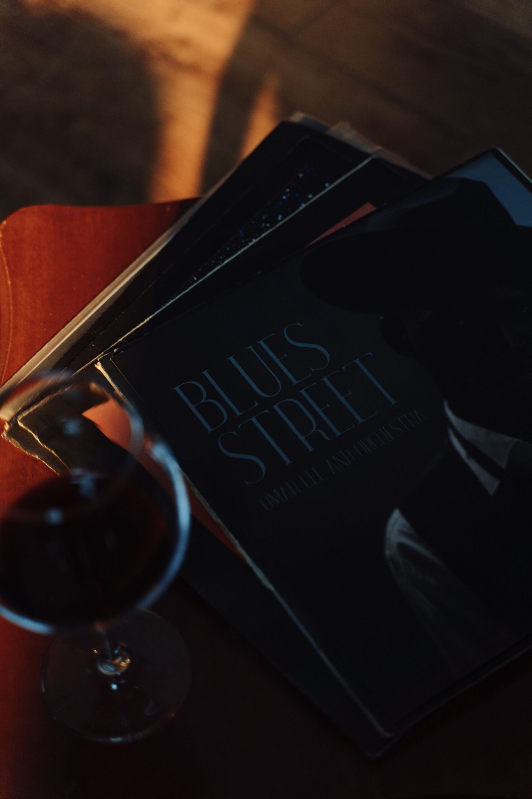 A glass of red wine sits on a table next to a stack of vinyl records, with Blues Street visible on top in dim, moody lighting.
