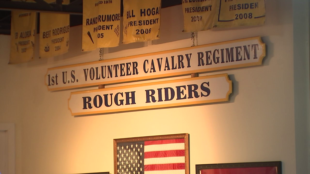 Wall display with signs reading 1st U.S. Volunteer Cavalry Regiment ROUGH RIDERS and parts of flags and yellow banners above with names and dates.