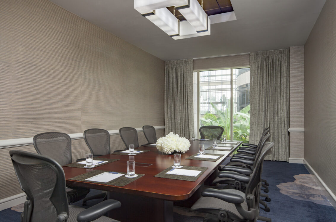 A modern conference room with a long table, ten chairs, notepads, glasses of water, and a bouquet of white flowers, lit by a large window.