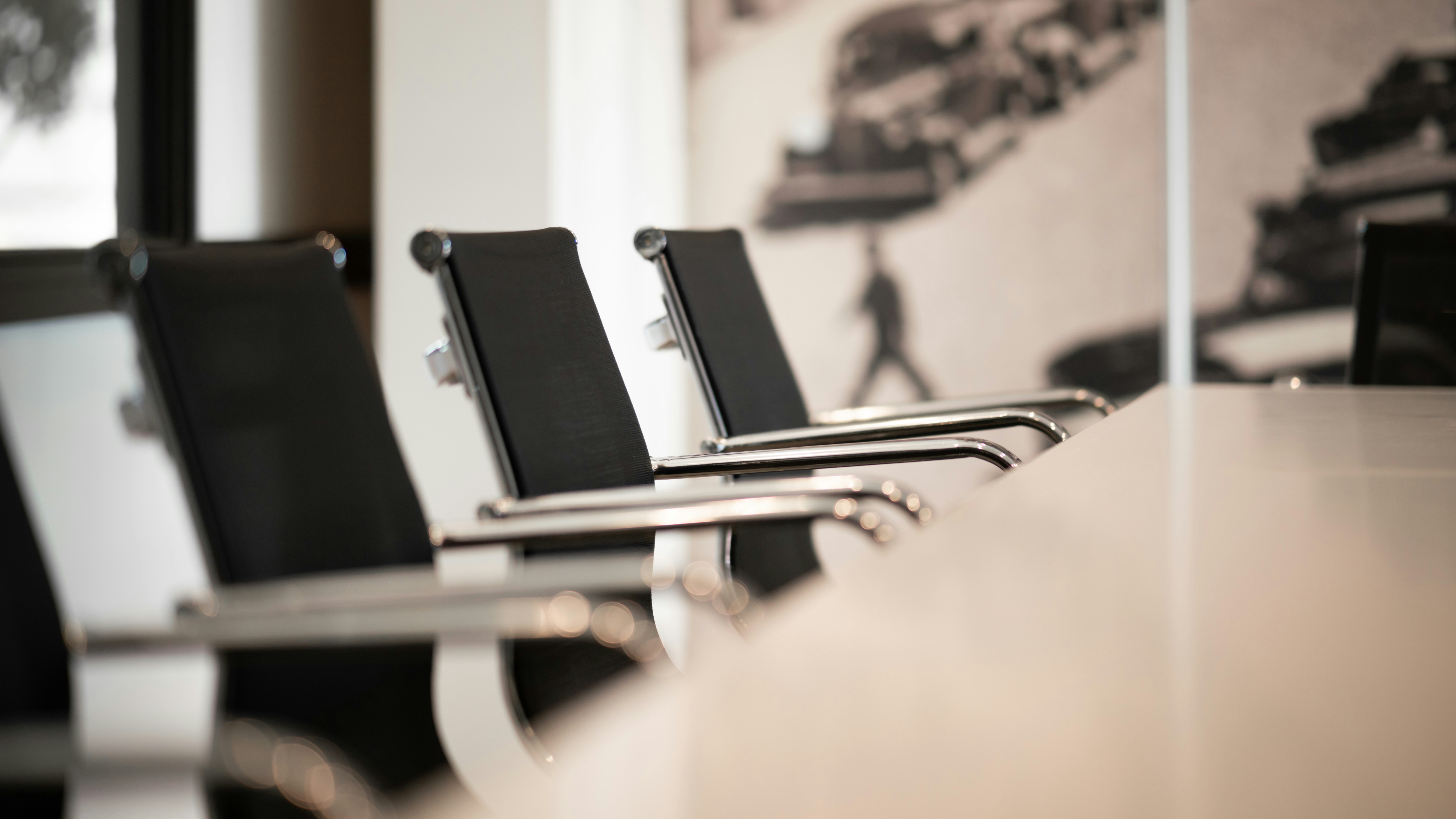 Four empty black office chairs are lined up at a conference table in a modern meeting room, with a blurry background.